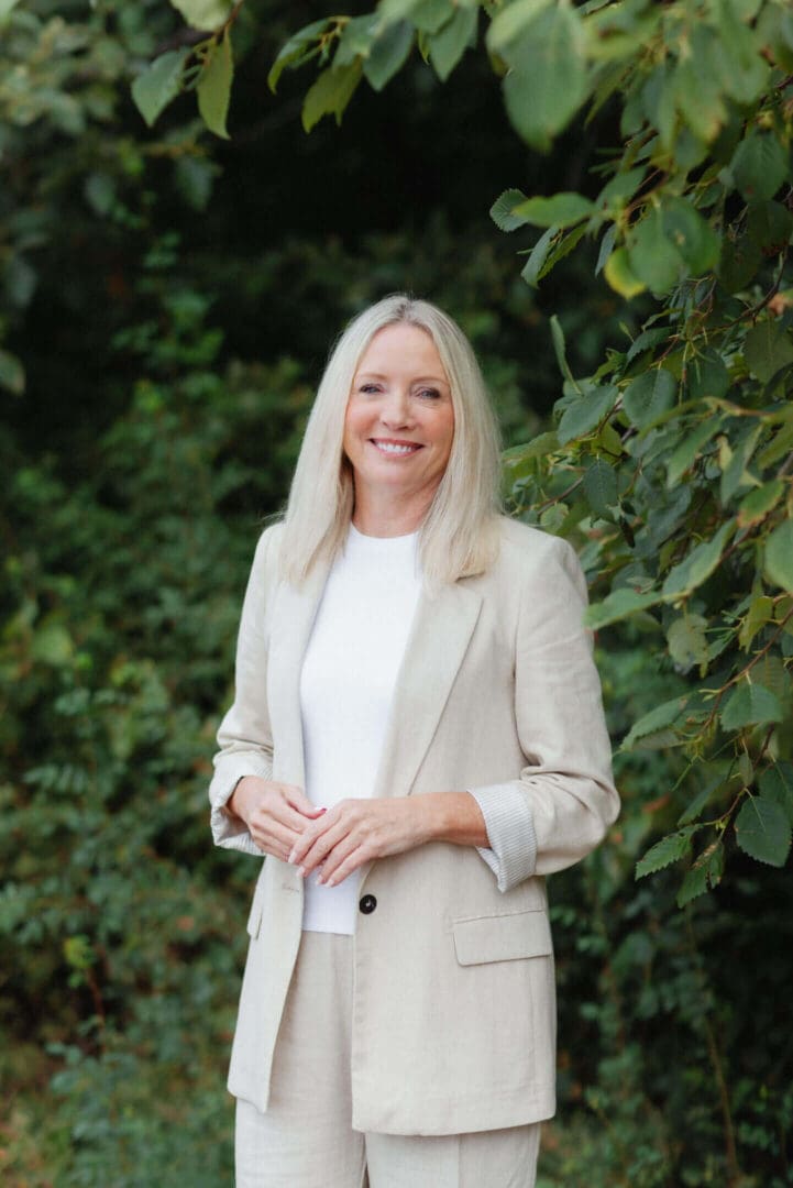 Smiling woman in beige blazer standing outdoors against green foliage.